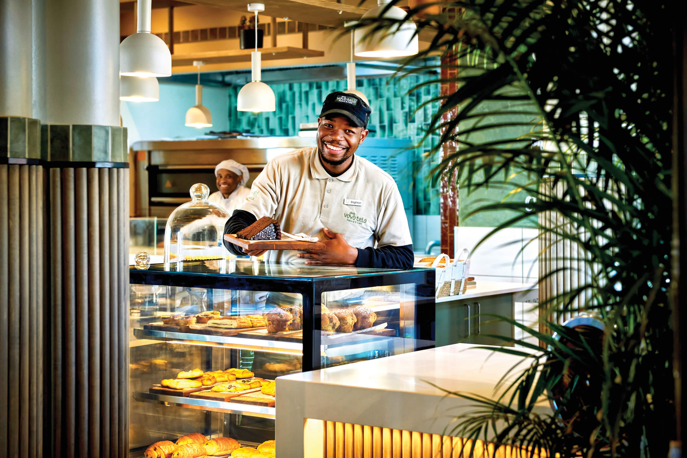 A smiling Vovo Telo baker holding a slice of cake at the pastry counter